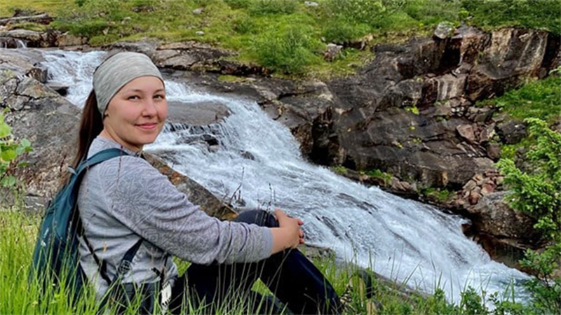 Bilde av mørkhåret jente i 20-årene ute i naturen som sitter ved siden av en bekk. 